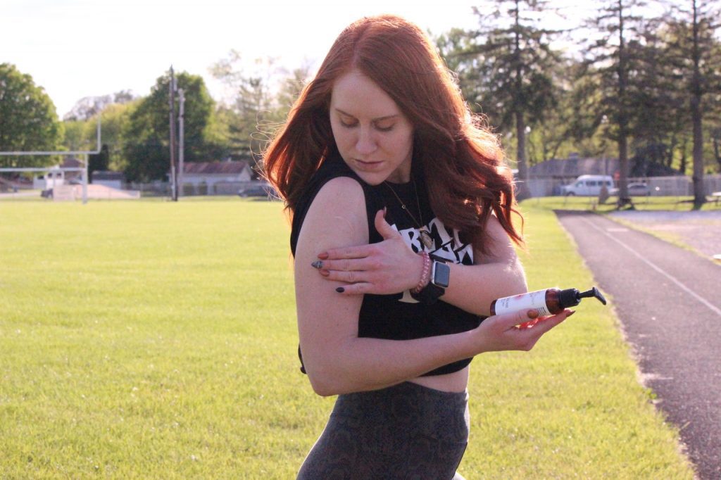 A young woman applies Nature's Candy organic CBD massage cream to her right arm in an open field. The woman is wearing a black tank top and gym sweat pants, and she has long brown hair. The field is green and lush, with a blue sky and white clouds in the background.